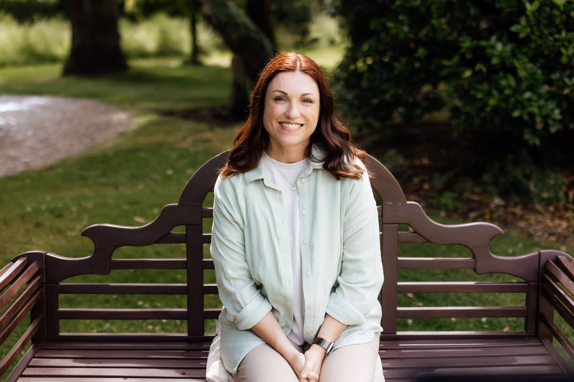 Smiling woman with auburn hair sitting on a wooden park bench wearing a light green shirt and beige pants.