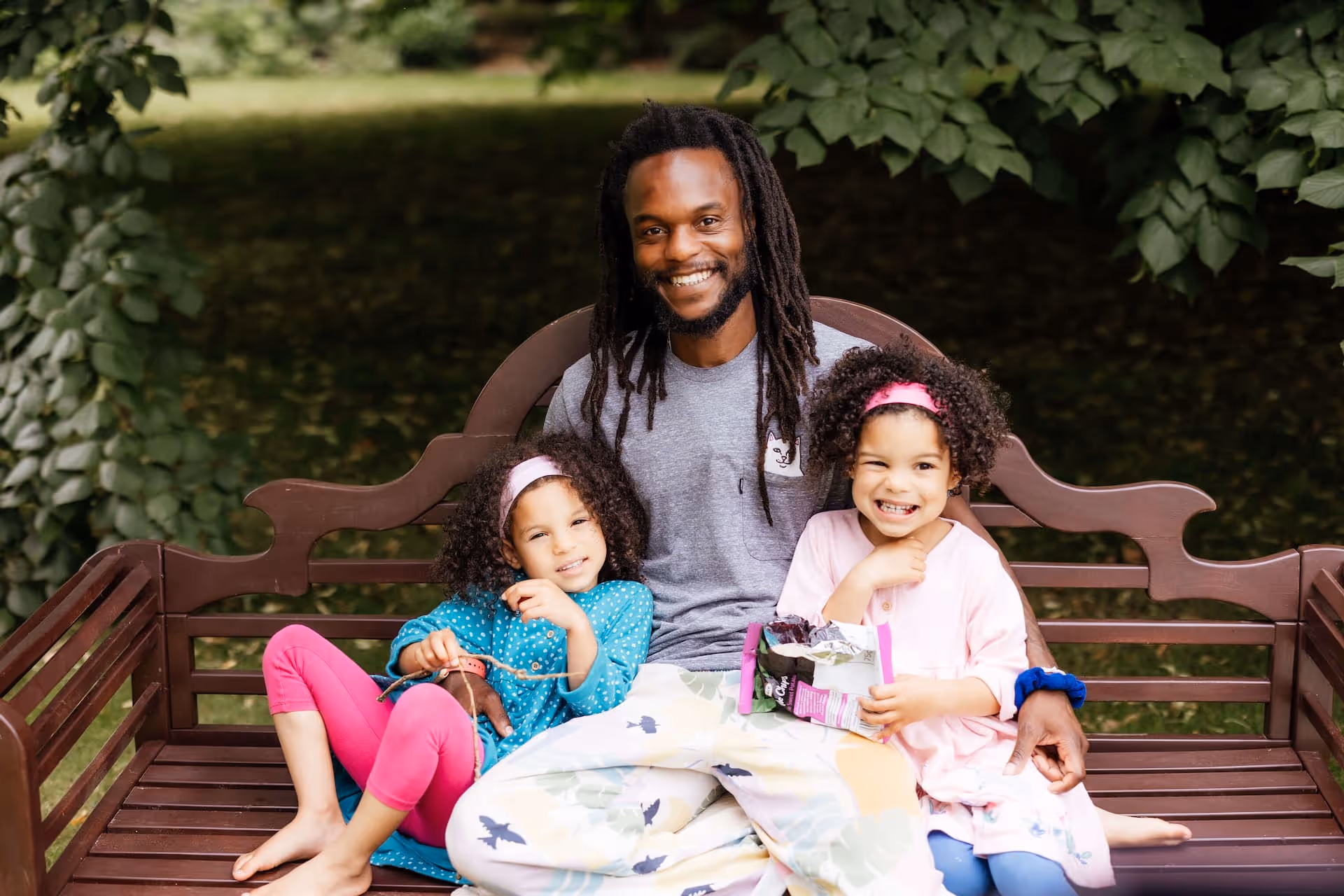 Smiling man with dreadlocks sitting on a wooden bench outdoors with two young girls, one holding a snack and the other wearing pink leggings.