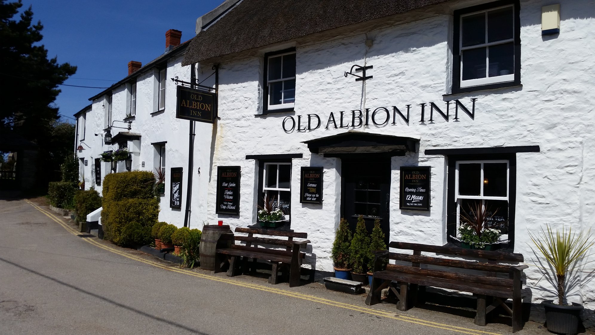 Old Albion Inn, a whitewashed building with thatched roof, black wooden benches, and signs on its facade along a quiet road.