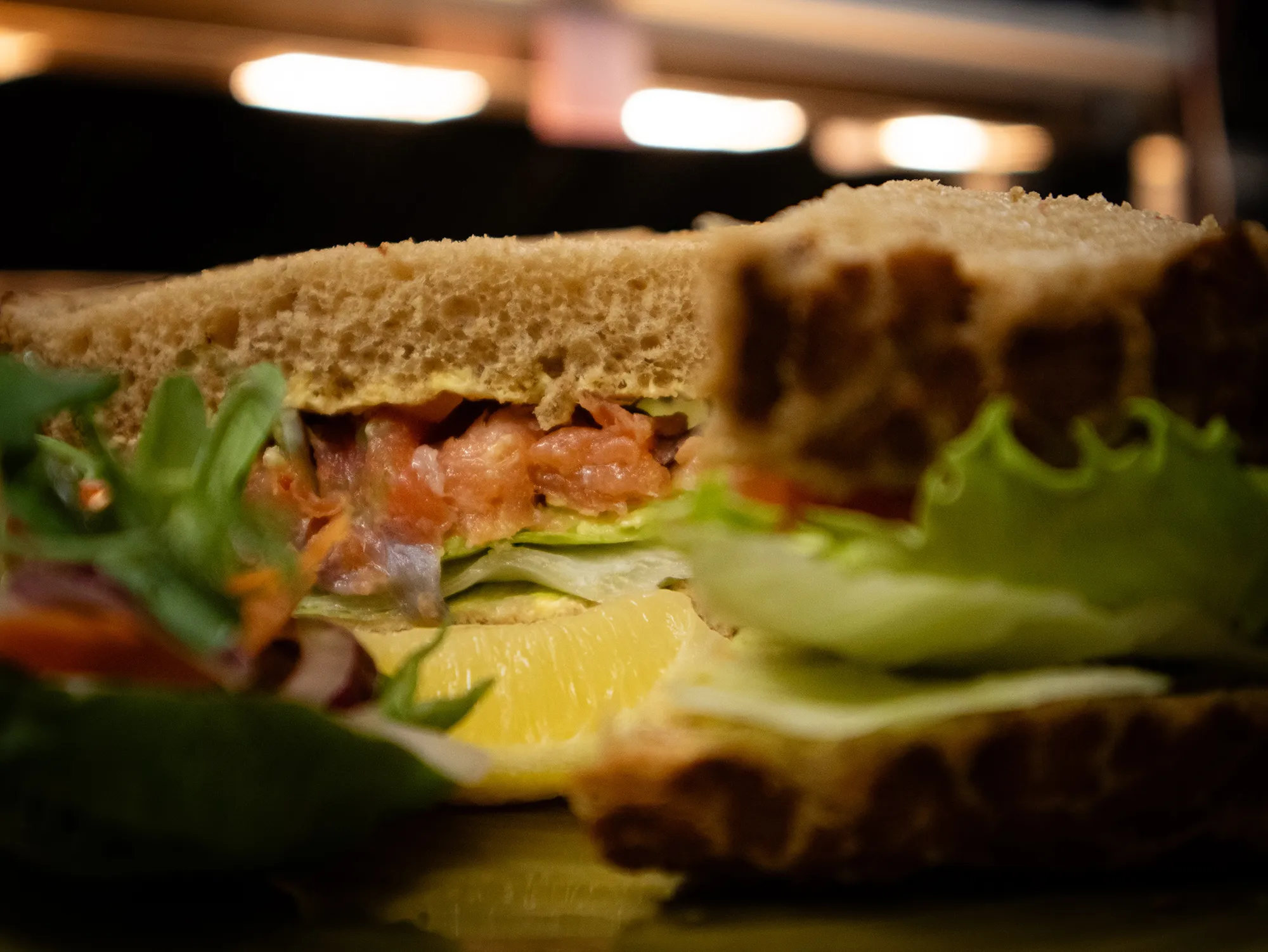 Close-up of a sandwich with brown bread, lettuce, slices of salmon, and a lemon wedge, with leafy greens on the side.