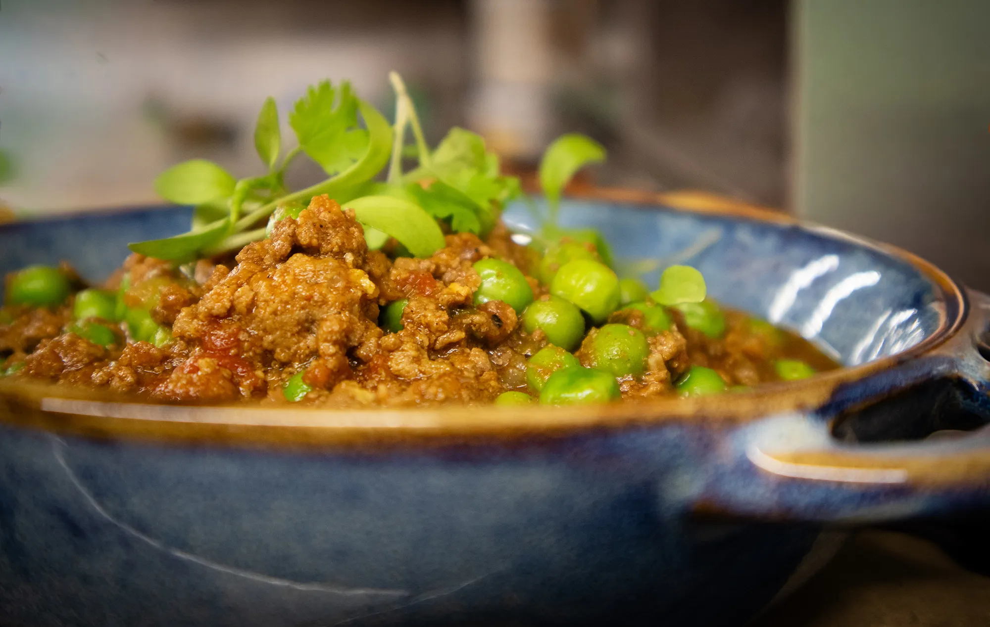 Close-up of minced meat curry with green peas and fresh herbs in a blue ceramic bowl.