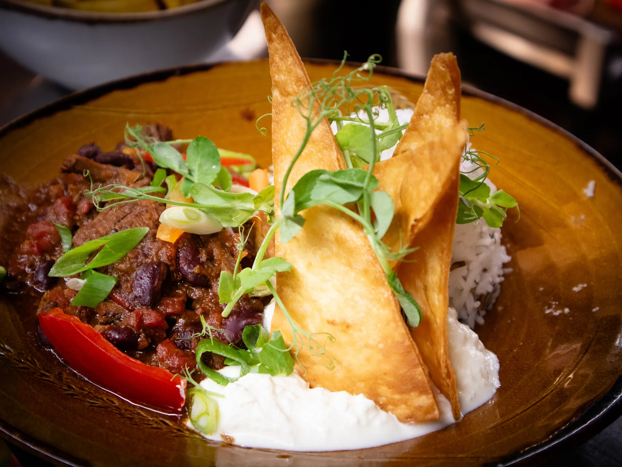 Plate with chili con carne garnished with green onions and a red pepper slice, white rice, two crispy triangular tortilla chips, sour cream, and fresh microgreens.