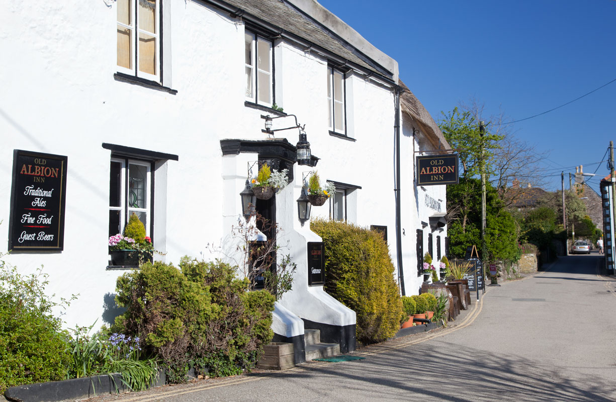 White traditional pub called Old Albion Inn with hanging flower baskets and black-framed windows along a quiet street under a clear blue sky.