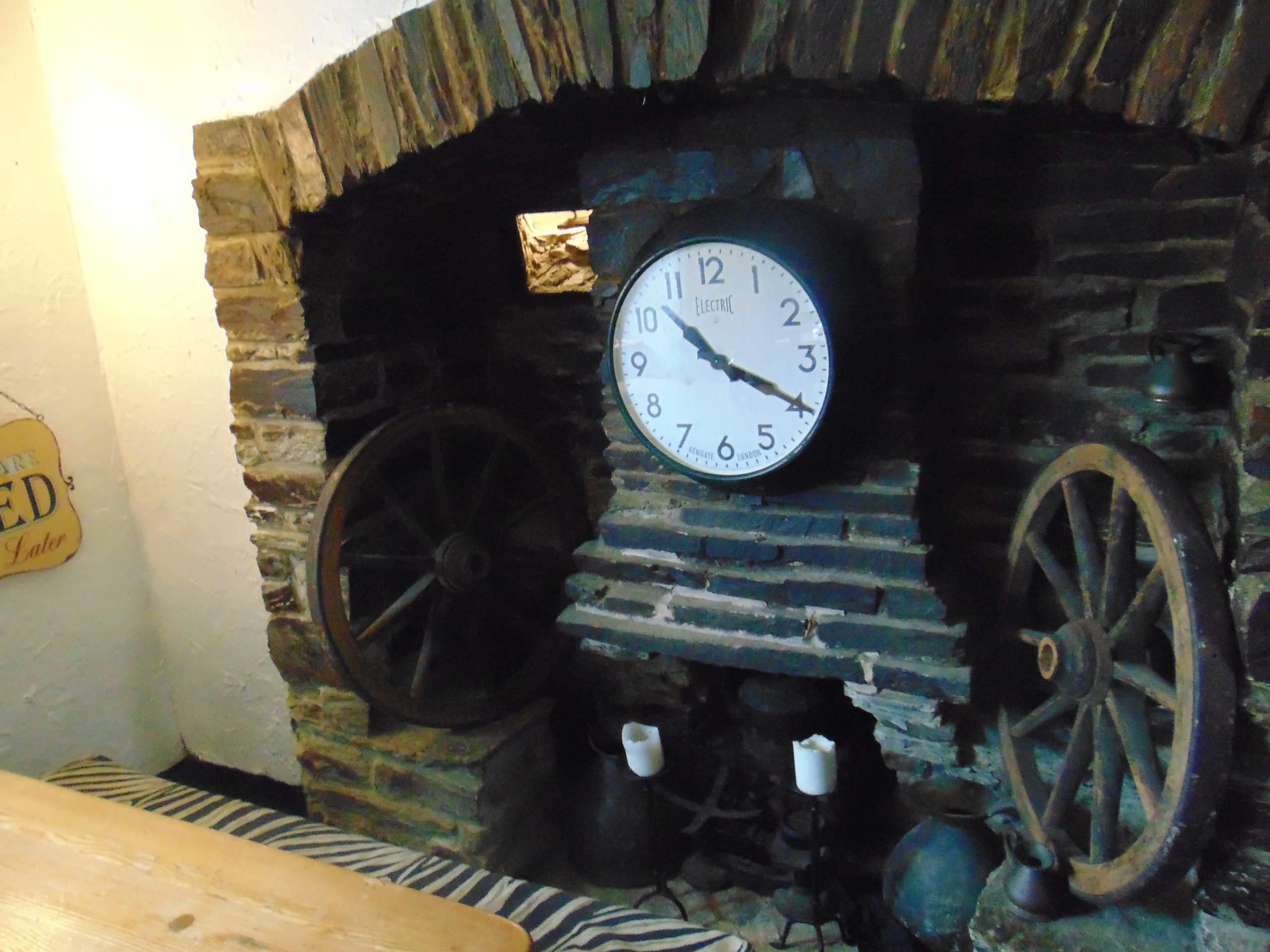 Rustic stone fireplace with a round white clock reading 10:20, flanked by two large wooden wagon wheels and old metal containers with two white candles in front.