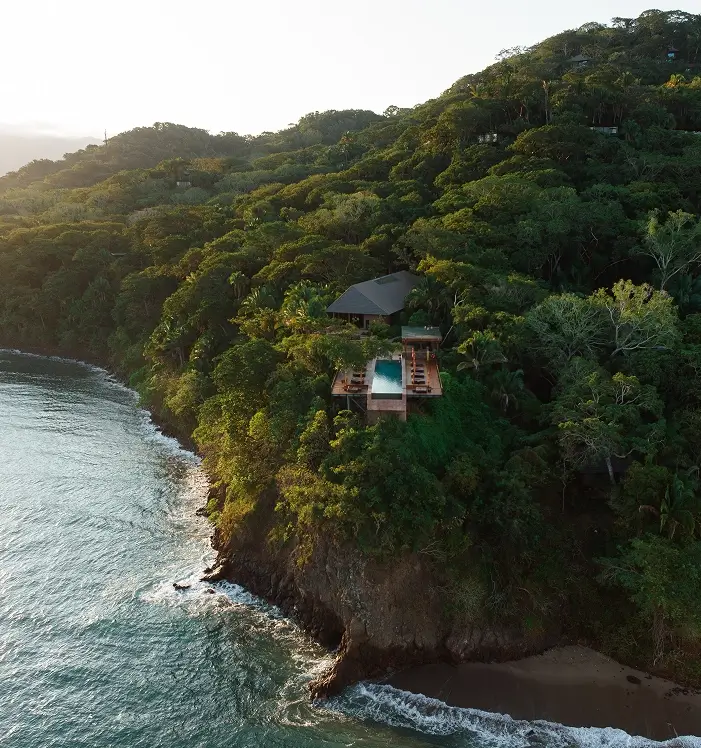 A private house with an infinity pool on a lush green cliff overlooking the ocean at sunset.