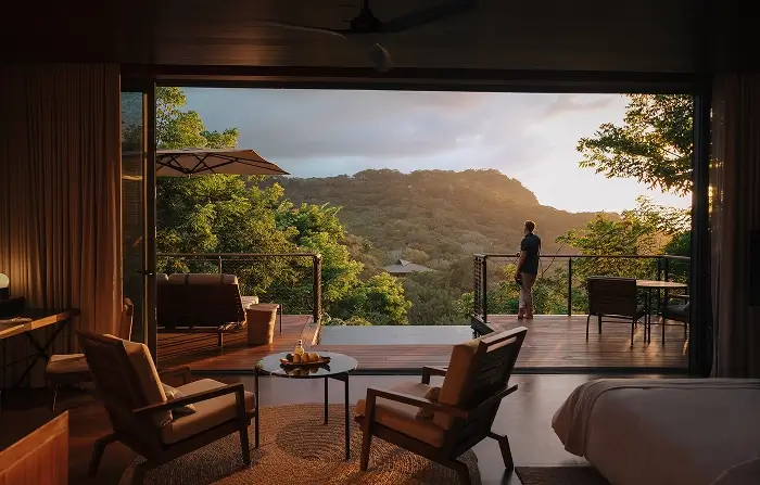 Hotel room with wooden chairs and table opening to balcony overlooking a person standing and a lush green mountain at sunset.