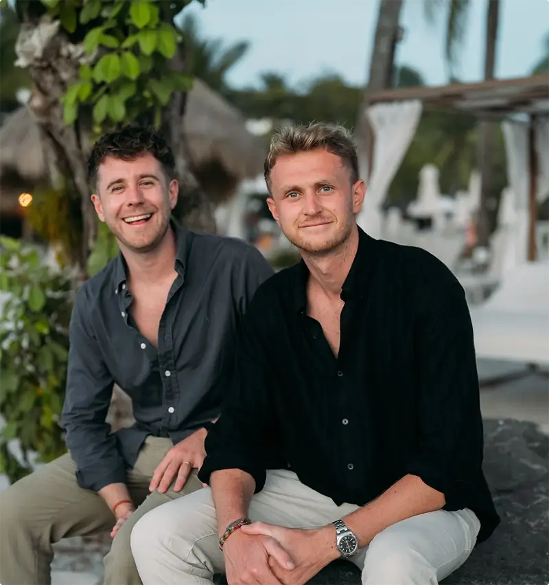 Two casually dressed men sitting outdoors near tropical plants and smiling at the camera.