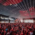 Large crowd at a concert or event with raised hands under a ceiling with red and white light patterns.