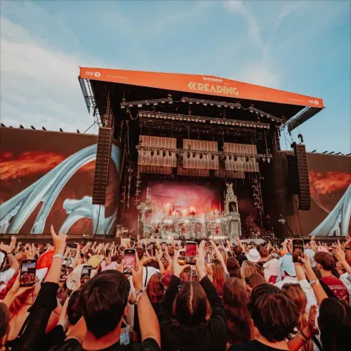 Crowd holding up phones and watching a large outdoor music festival stage with 'Reading' banner and elaborate set design.