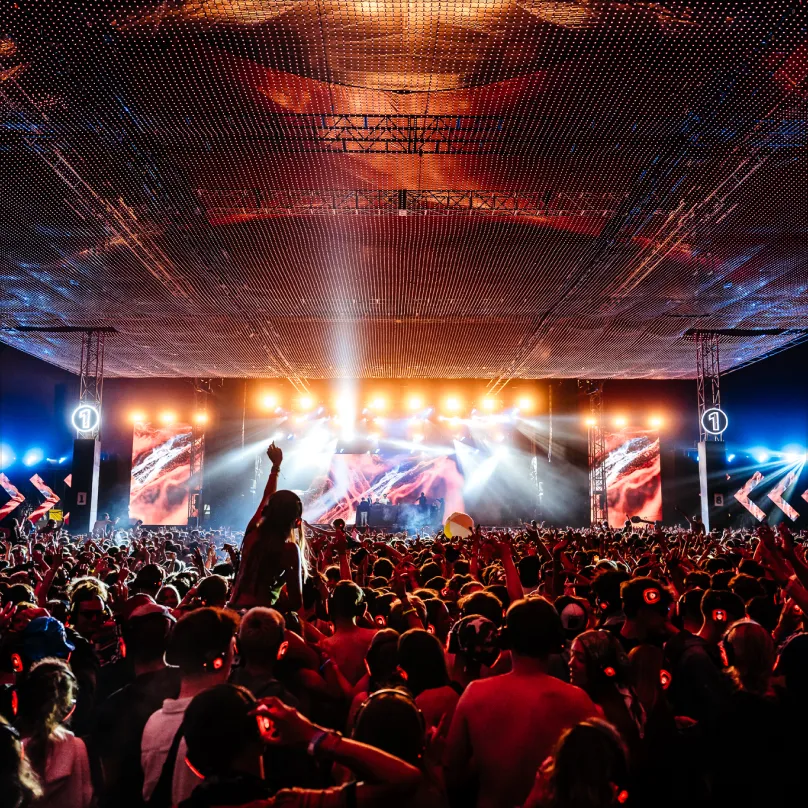 Large crowd wearing headphones enjoying a vibrant concert with bright stage lights and visual screens under a textured ceiling.