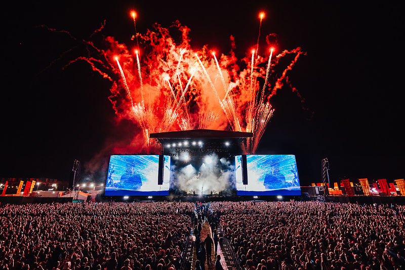 A large crowd of people in front of the main stage with fireworks at night.