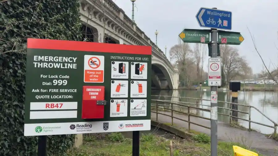 A safety sign by a river shows instructions for using an emergency throwline, with a bridge in the background.