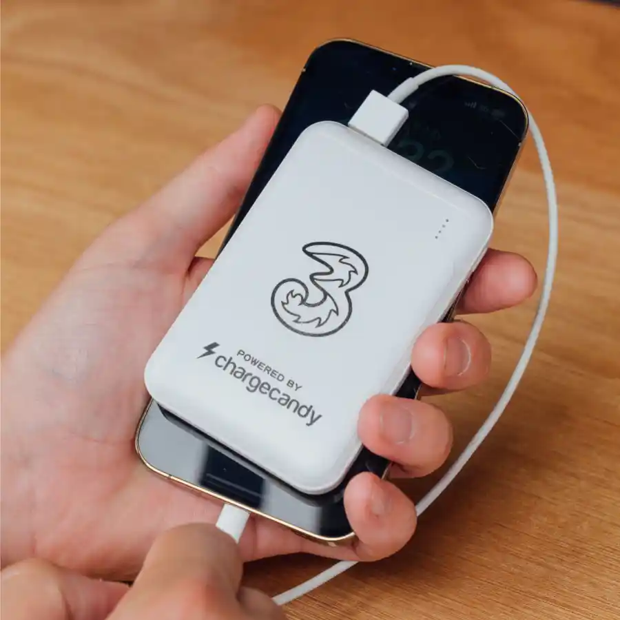 A hand holds a smartphone connected to a white power bank featuring the number "3" and "powered by chargecandy" logo, on a wooden table.
