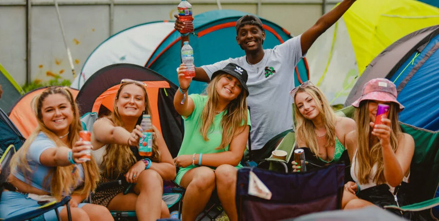 Group of friends smiling and raising drinks while relaxing in a campsite surrounded by colorful tents.