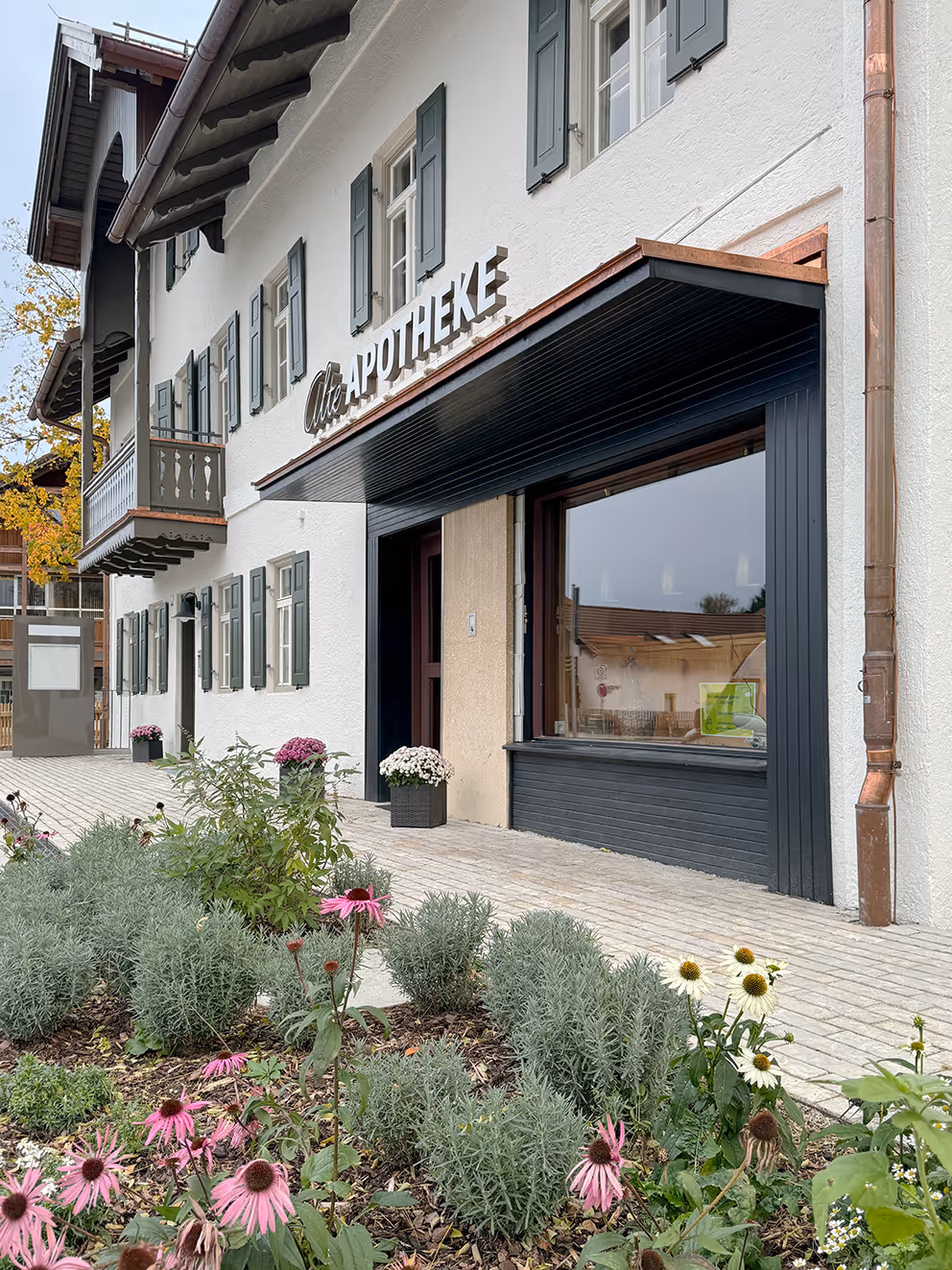 Außenansicht einer weißen Apotheke mit grünen Fensterläden, Balkon und Blumenbeeten im Vordergrund.