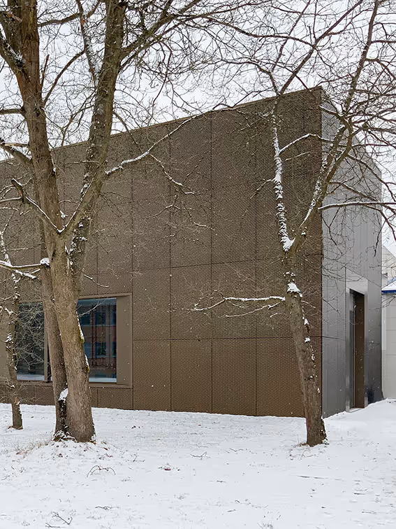 Braunes modernes Gebäude mit großen Fenstern, im Vordergrund Bäume mit leichtem Schneebelag und schneebedeckter Boden.
