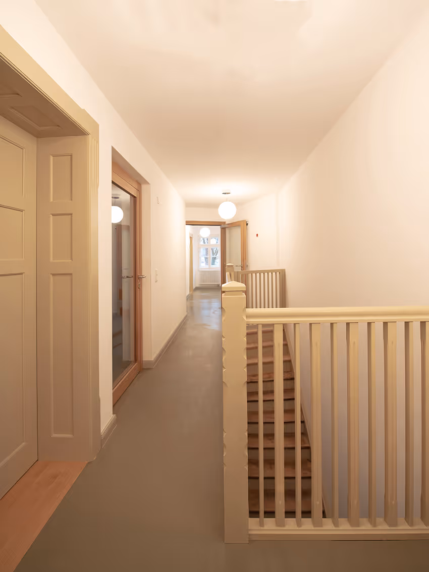 Heller Flur mit beige gestrichenem Treppengeländer, geschlossenen Türen und runden Deckenlampen.