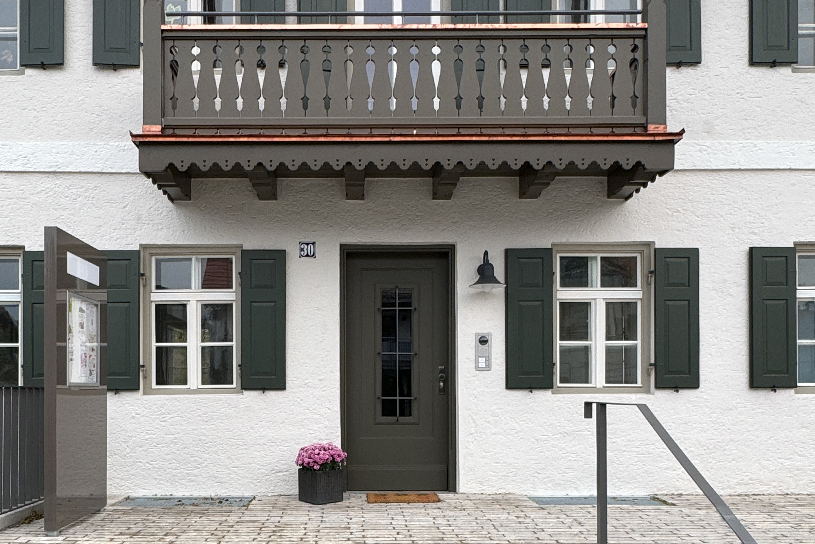 Fassade eines weißen Gebäudes mit dunkelgrüner Tür und Fensterläden, darüber ein Balkon mit Holzgeländer und Topf mit rosa Blumen neben der Eingangstür.