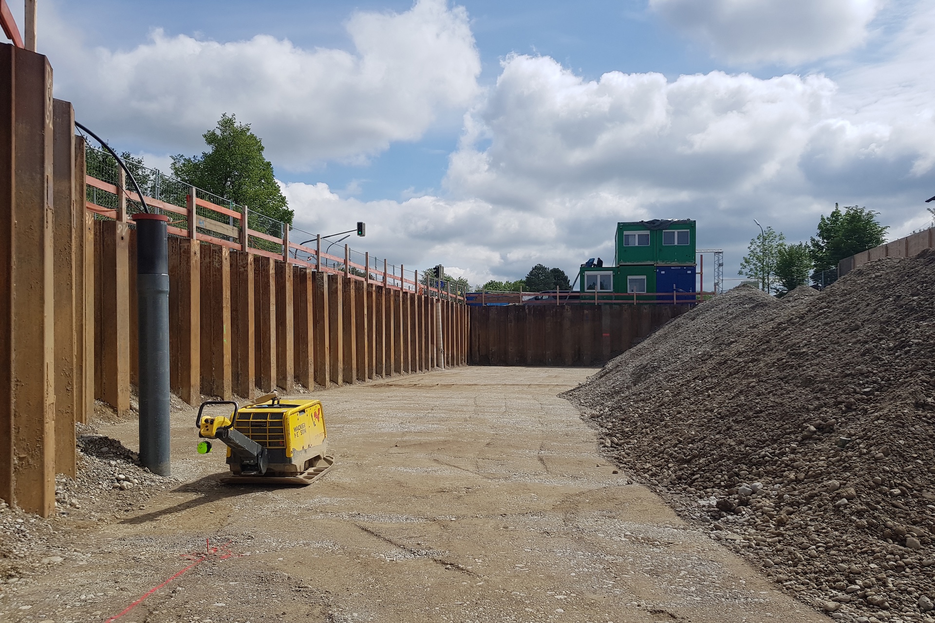 Baustelle mit einem großen Erdloch, großen Erdhaufen rechts, einem gelben Verdichtergerät in der Mitte und grünen Baucontainern im Hintergrund.