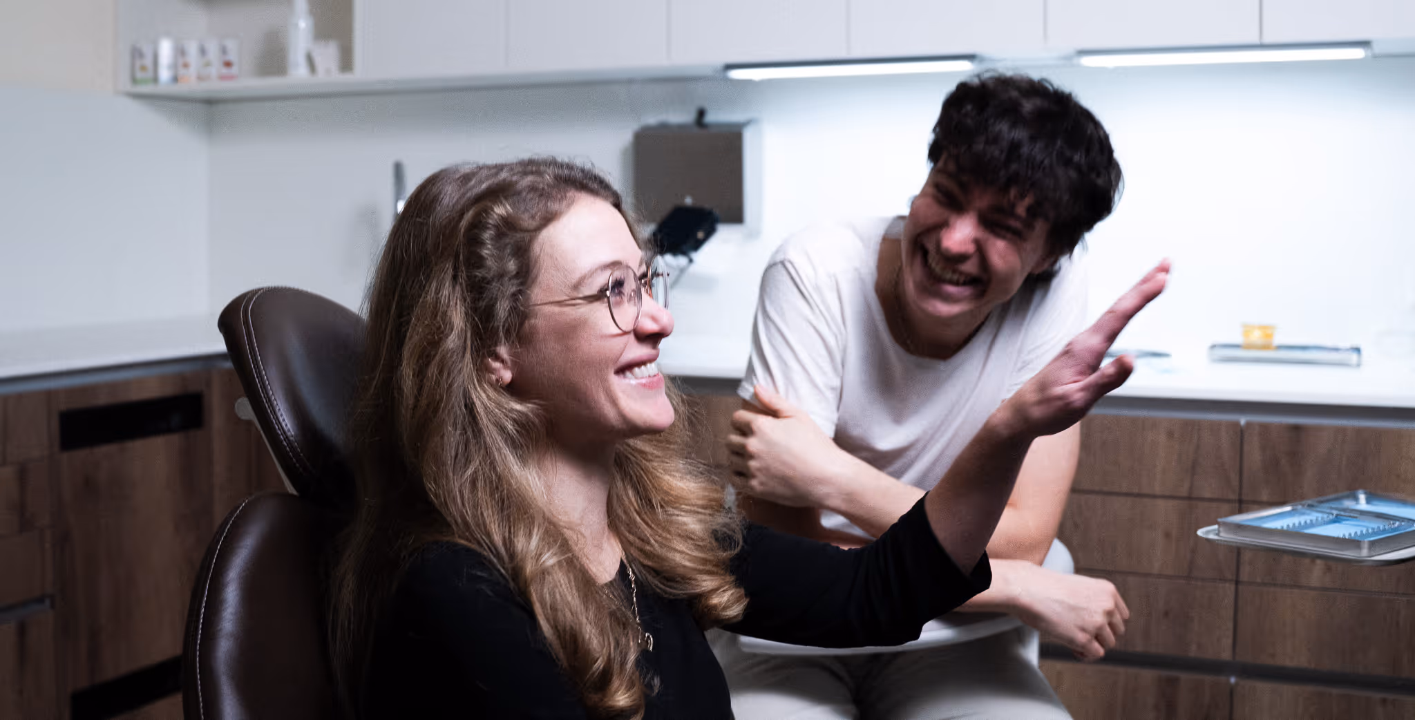 Smiling woman with glasses sitting in a chair waving her hand, while a laughing man sits behind her in a modern kitchen.