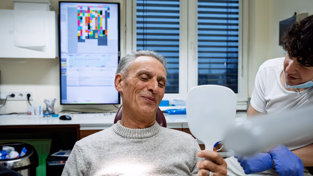 Smiling elderly man looking into a hand mirror while a medical professional in gloves stands beside him in a clinical setting.