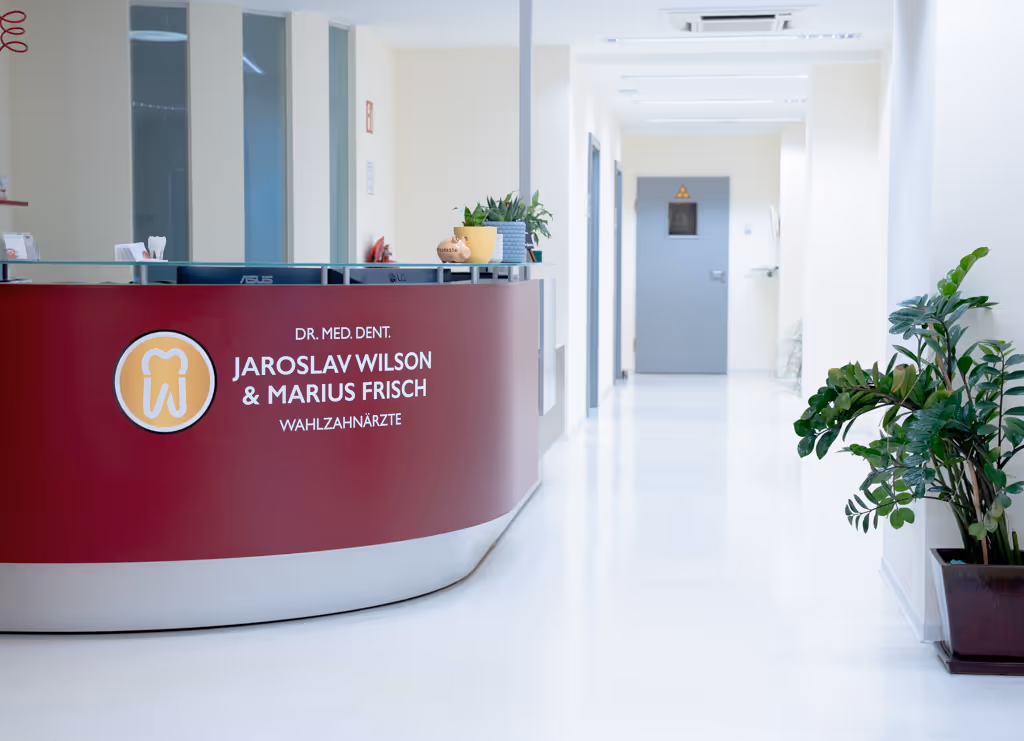 Modern dental clinic reception area with a red curved desk displaying names of dentists Jaroslav Wilson and Marius Frisch, with plants on the desk and a hallway in the background.