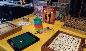 A collection of board games including Dobble, 7 Wonders, Codenames, Junk Art, Othello, Pick Up Sticks, and a rainbow slinky, arranged ready to play on a yellow foyer table