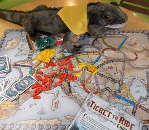 The board game Ticket to Ride on a table, showing a map board and many small plastic game pieces and cards, with a green plush iguana soft toy next to it