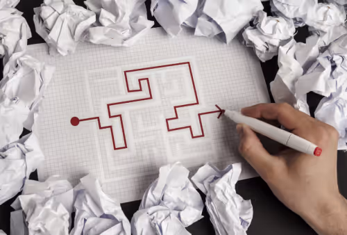 a paper maze puzzle with the route drawn in red pen by a hand, surrounded by crumpled paper