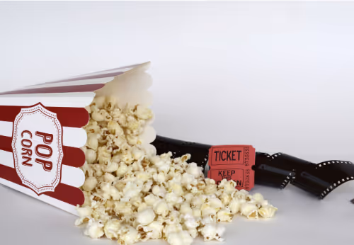 A red and white striped tub of movie popcorn on its side next to a reel of film and two red film tickets