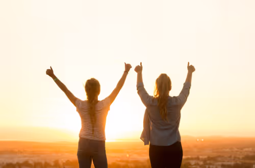 two people with their backs to the camera doing double thumbs up against the sunset