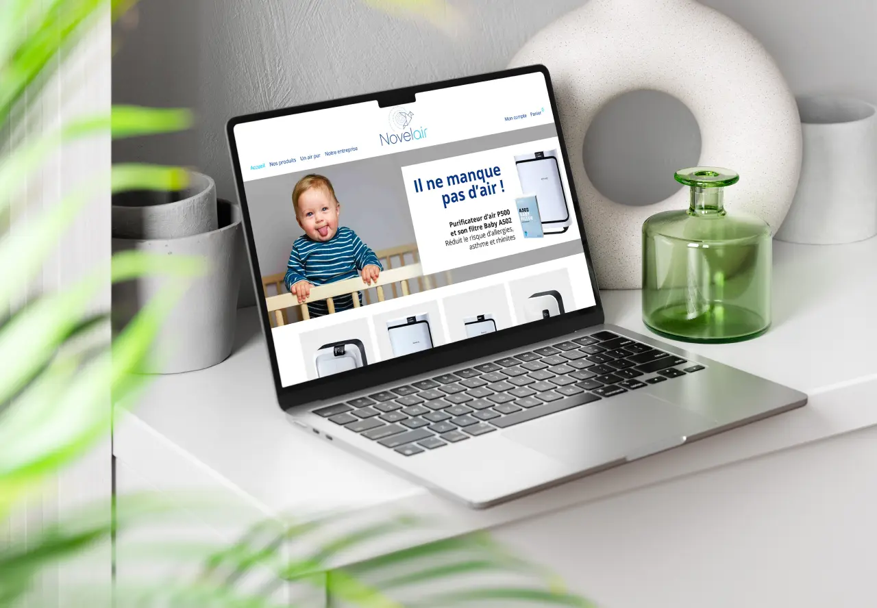 Laptop affichant le site Novelair avec un bébé et le message Il ne manque pas d'air, entouré d'une plante verte, d'une tasse et d'un vase