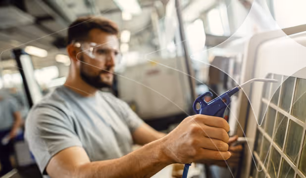 Man wearing safety glasses using a blue air blower to clean a tiled surface in an industrial or workshop setting.