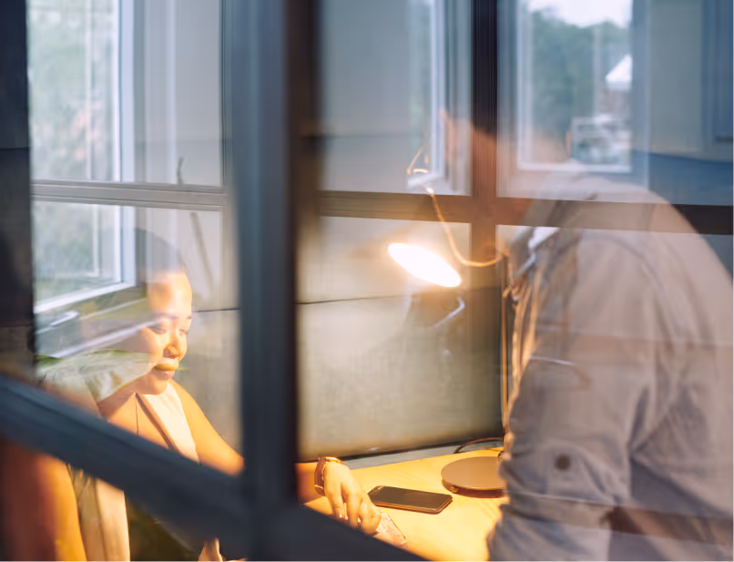 Two people working together at a desk by a window, viewed through glass panels with reflections.