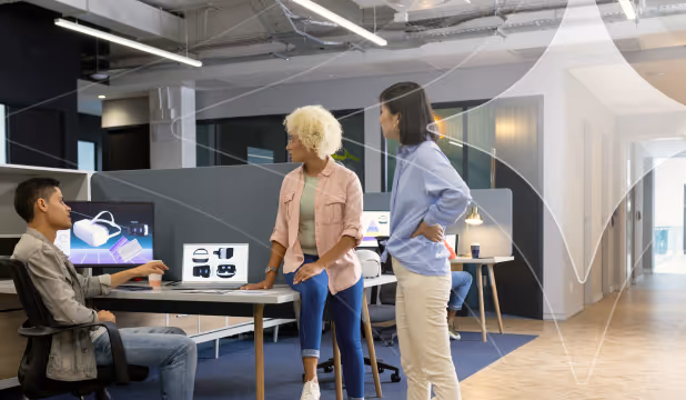 Three coworkers discussing VR headset images on computers in an office meeting area.
