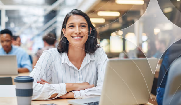 Smiling woman in white shirt sitting at desk with laptop and coffee cup in a busy office.