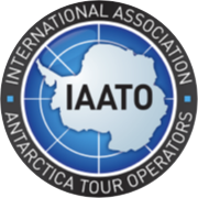 Logo of the International Association of Antarctica Tour Operators (IAATO), featuring a stylized white map of Antarctica centered on a blue globe grid. The acronym "IAATO" is prominently displayed across the continent image. The circular emblem is bordered by a dark ring with the text "INTERNATIONAL ASSOCIATION" at the top and "ANTARCTICA TOUR OPERATORS" at the bottom in white capital letters. Keywords: IAATO logo, Antarctica tourism, polar expedition regulation, sustainable travel, Antarctic operators, environmental protection.