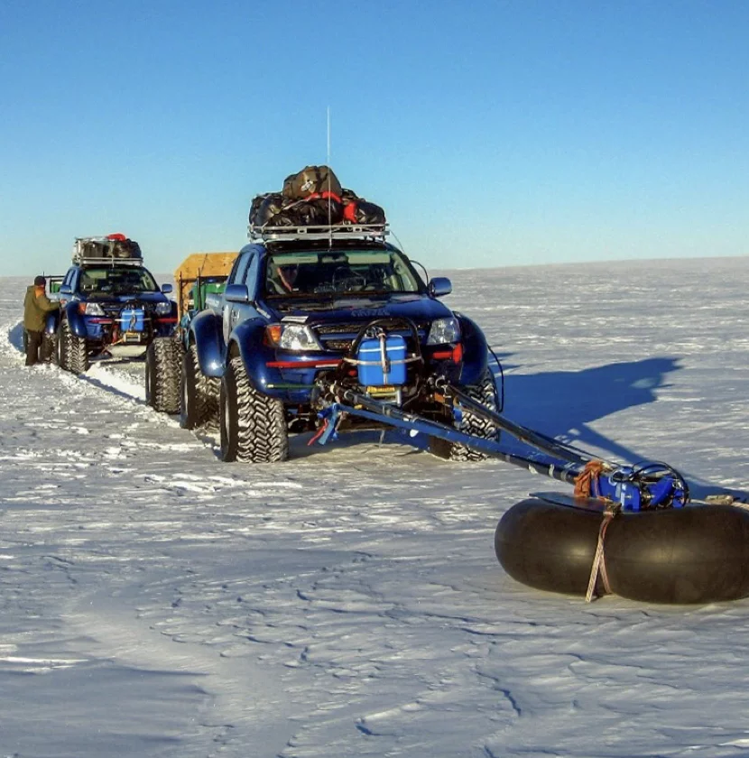 Two blue off-road vehicles equipped with gear on their roofs driving on a snowy, icy terrain with a ground penetrating radar equipment attached to the front vehicle.