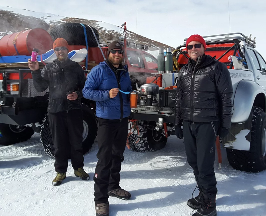 Three people wearing winter clothing stand on snow next to a truck loaded with large barrels and equipment, with steam rising from a cooking pot.