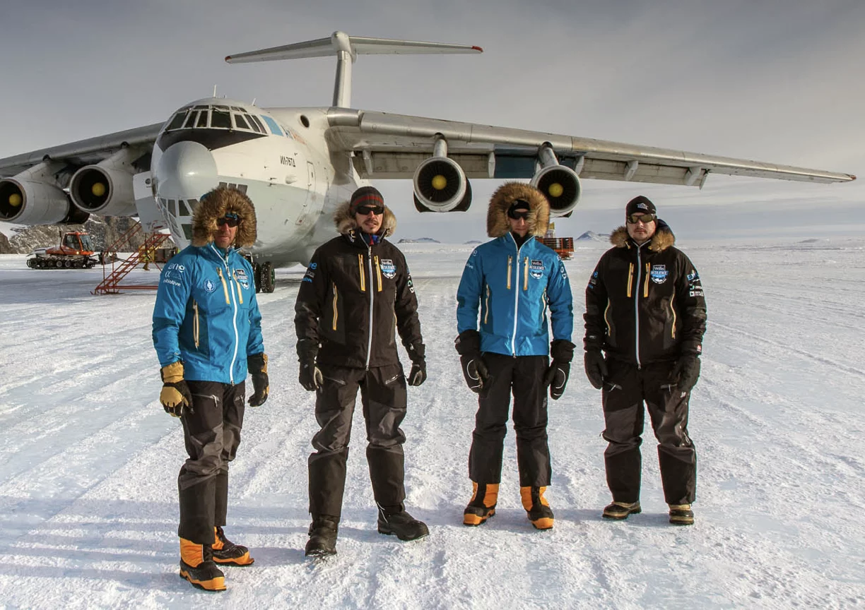 Four people in cold-weather gear standing on snow in front of a large cargo plane with four engines.