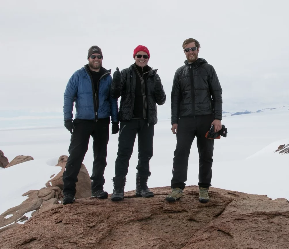 Three men dressed in winter gear standing on a rocky surface with snow-covered mountains in the background.