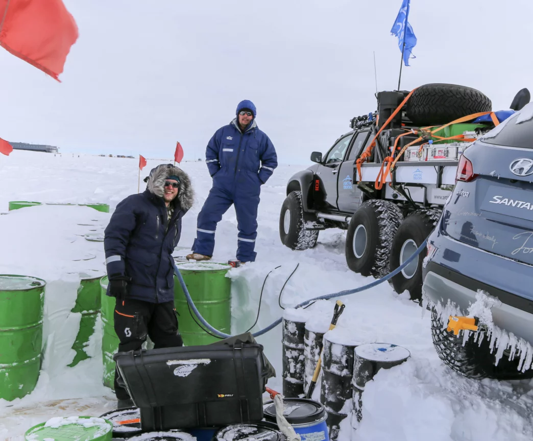 Two people in heavy winter clothing standing by green barrels in a snowy landscape with two off-road vehicles nearby.