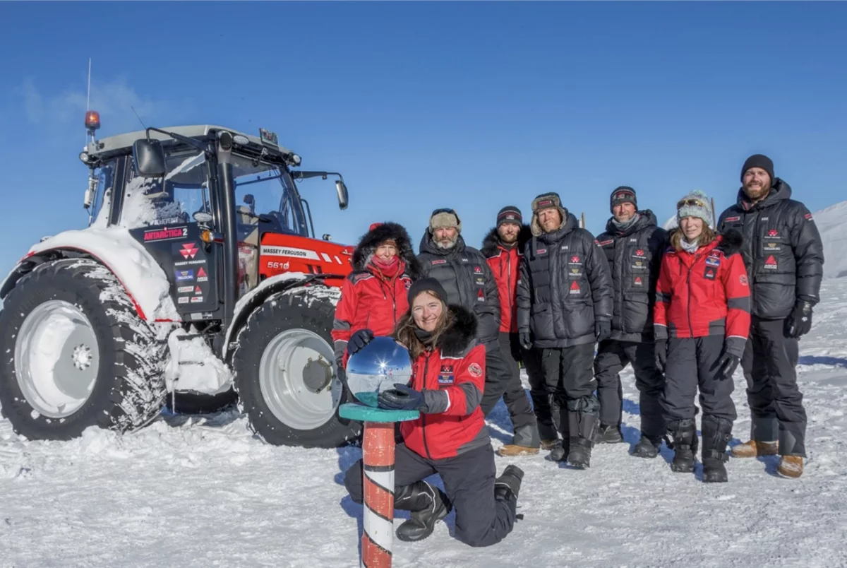 Group of nine people in winter gear posing on snow beside a red Massey Ferguson tractor under clear blue sky.