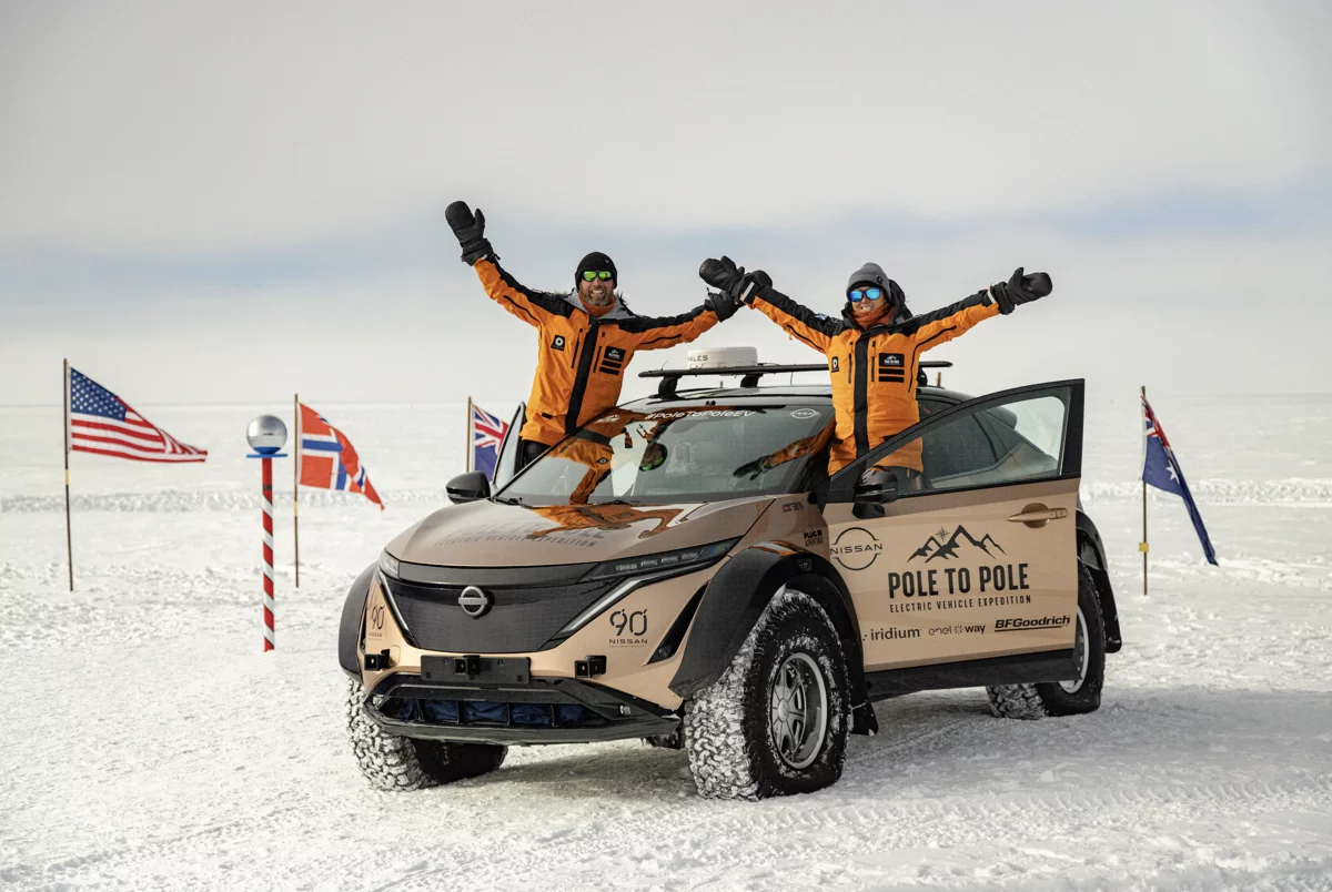 Two people in orange jackets with raised arms standing beside an electric Nissan vehicle in snowy terrain with several national flags in the background.