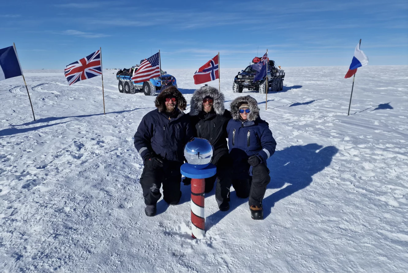 Three people in winter jackets kneeling on snow around a red, white, and blue striped pole with a silver sphere on top, with flags and off-road vehicles in the background on a snowy plain under a clear blue sky.