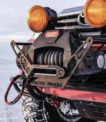 Front of an Arctic Trucks vehicle showing a COMEUP winch with visible steel cable and amber off-road lights, parked on snowy terrain.