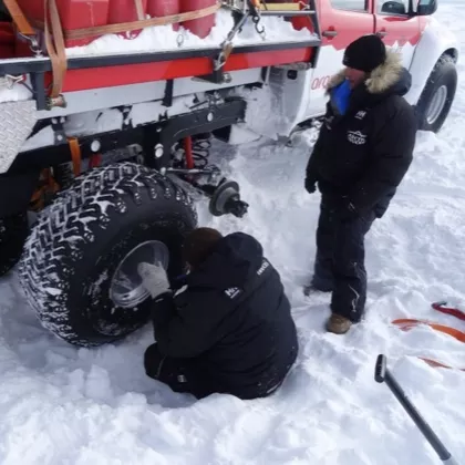 Two people in winter clothes working on a large tire of a snow-covered vehicle in a snowy environment.