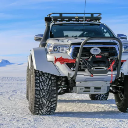 Off-road truck with large tires and winch on snowy terrain with mountains under blue sky.