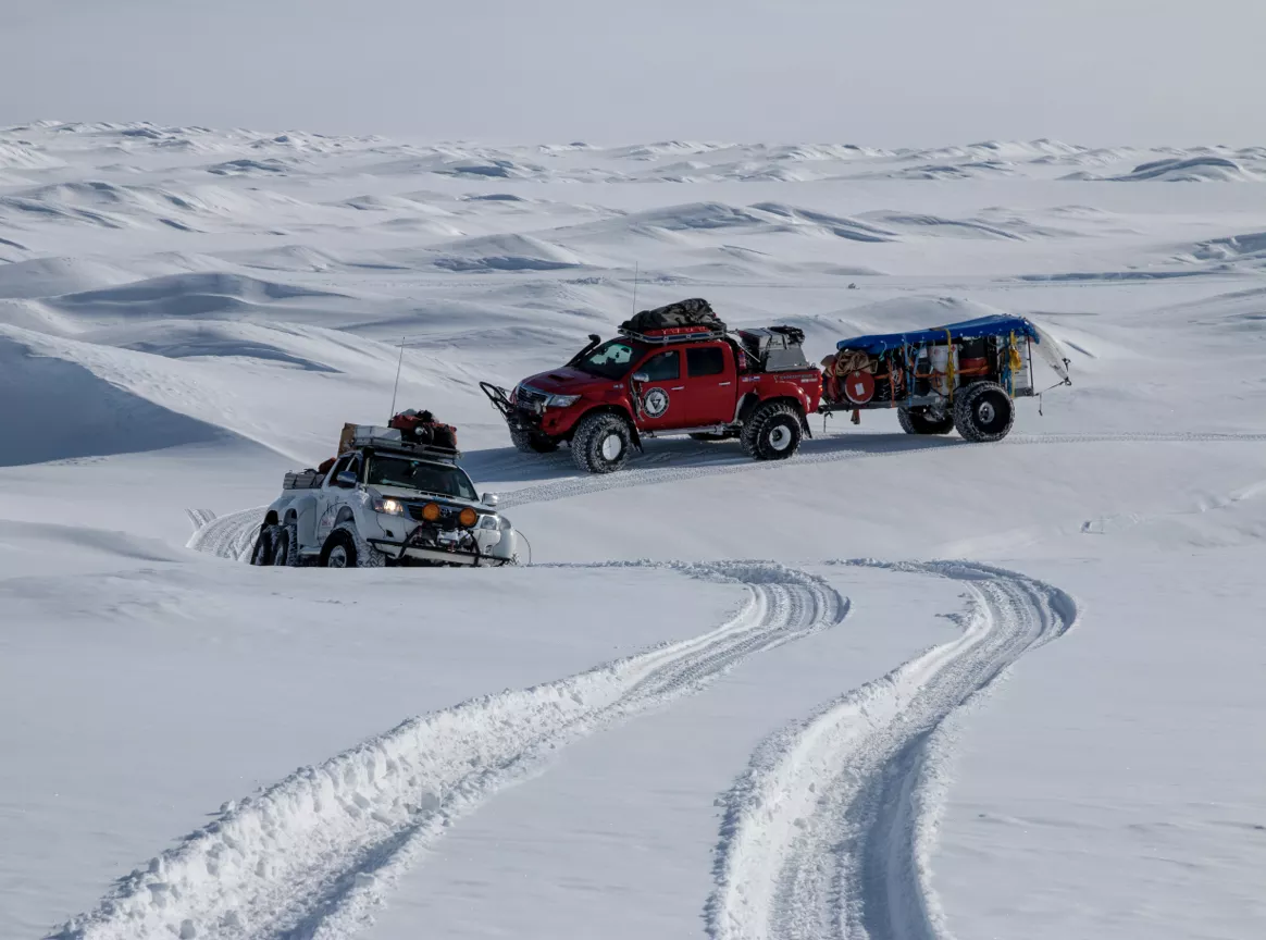 Two rugged off-road trucks, one red towing a trailer and one white, driving through a snowy landscape with tire tracks in the snow.