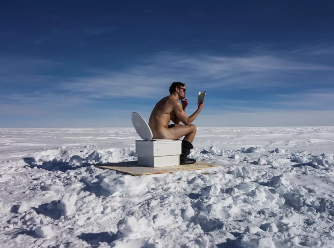 Naked man wearing sunglasses and boots sits on a white wooden box with a toilet seat lid, reading a book in a vast snowy landscape under a clear blue sky.